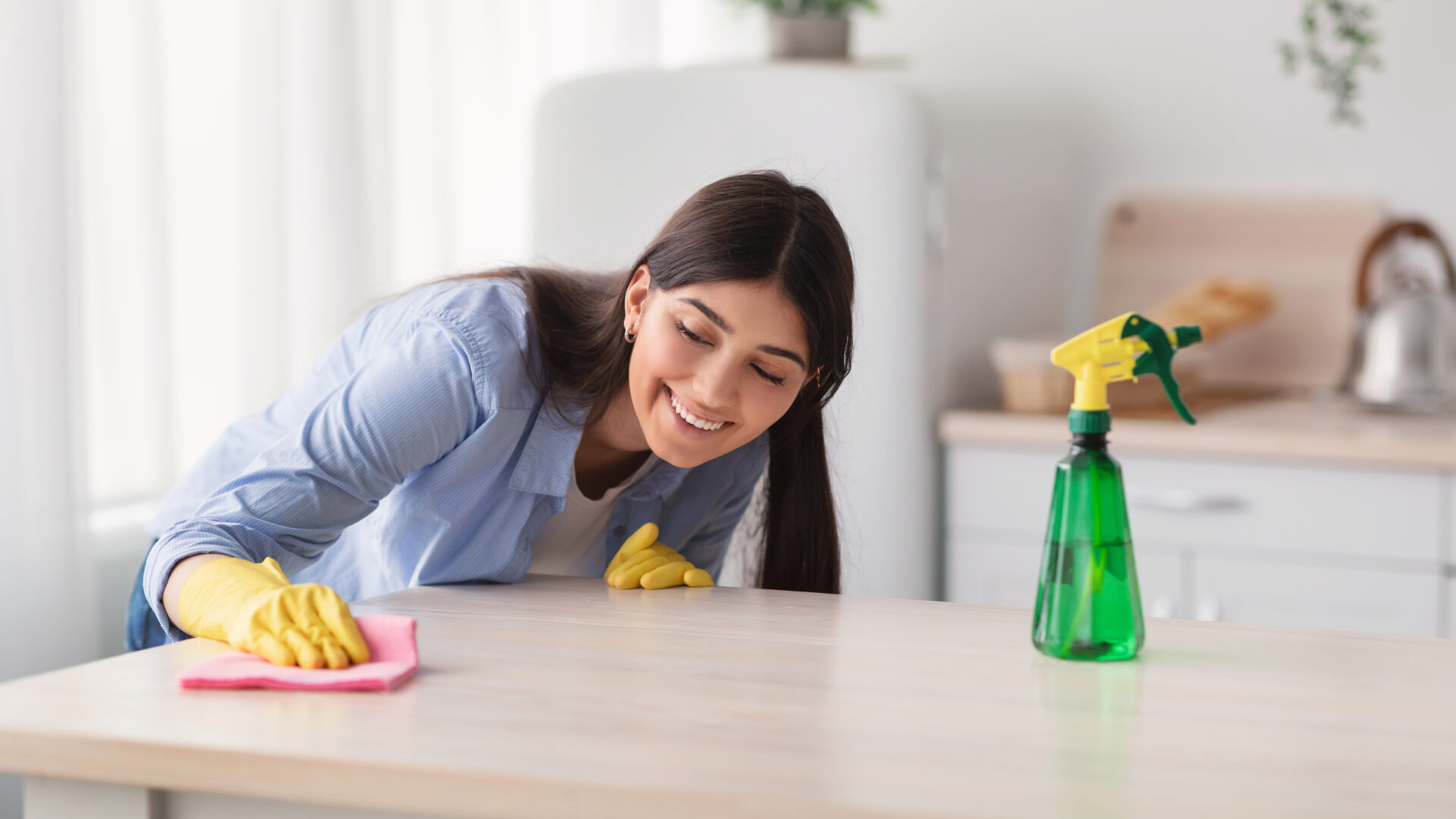 Portrait of happy young housewife in yellow rubber gloves cleaning dining table at kitchen. Positive millennial maid keeping her home tidy and clean, wiping surface with microfiber cloth
