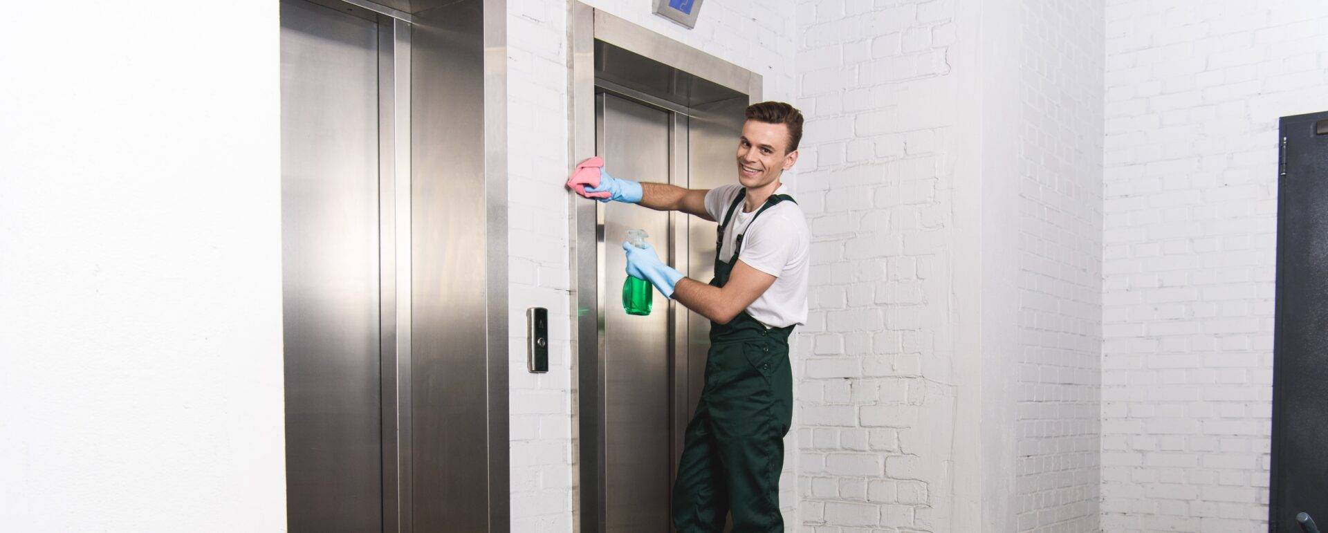handsome young janitor cleaning elevator and smiling at camera